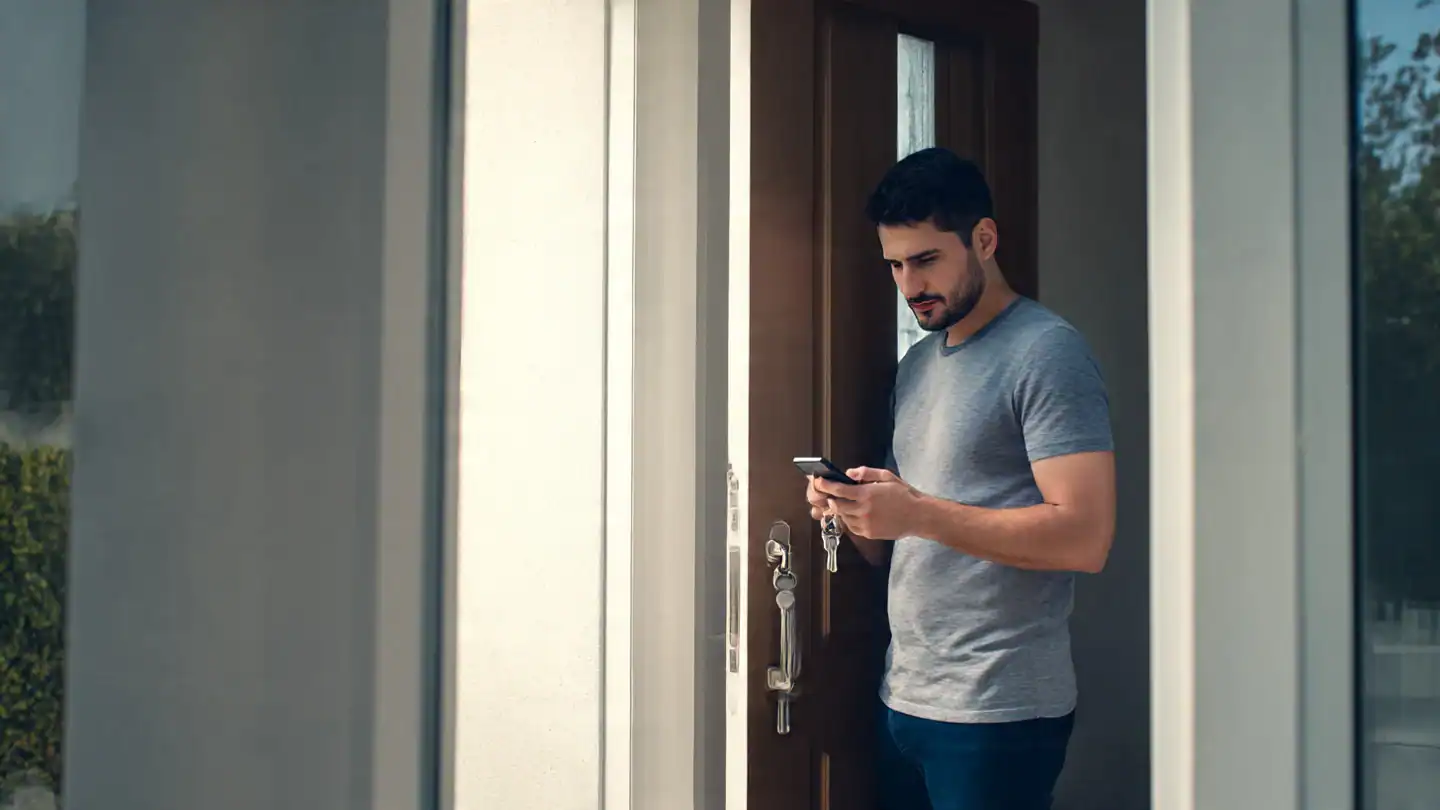 A man standing at his front door holding a smartphone and a metal key, looking slightly frustrated — symbolizing the contrast between modern smart home technology and old-fashioned door locks. smart locks that work with Apple HomeKit